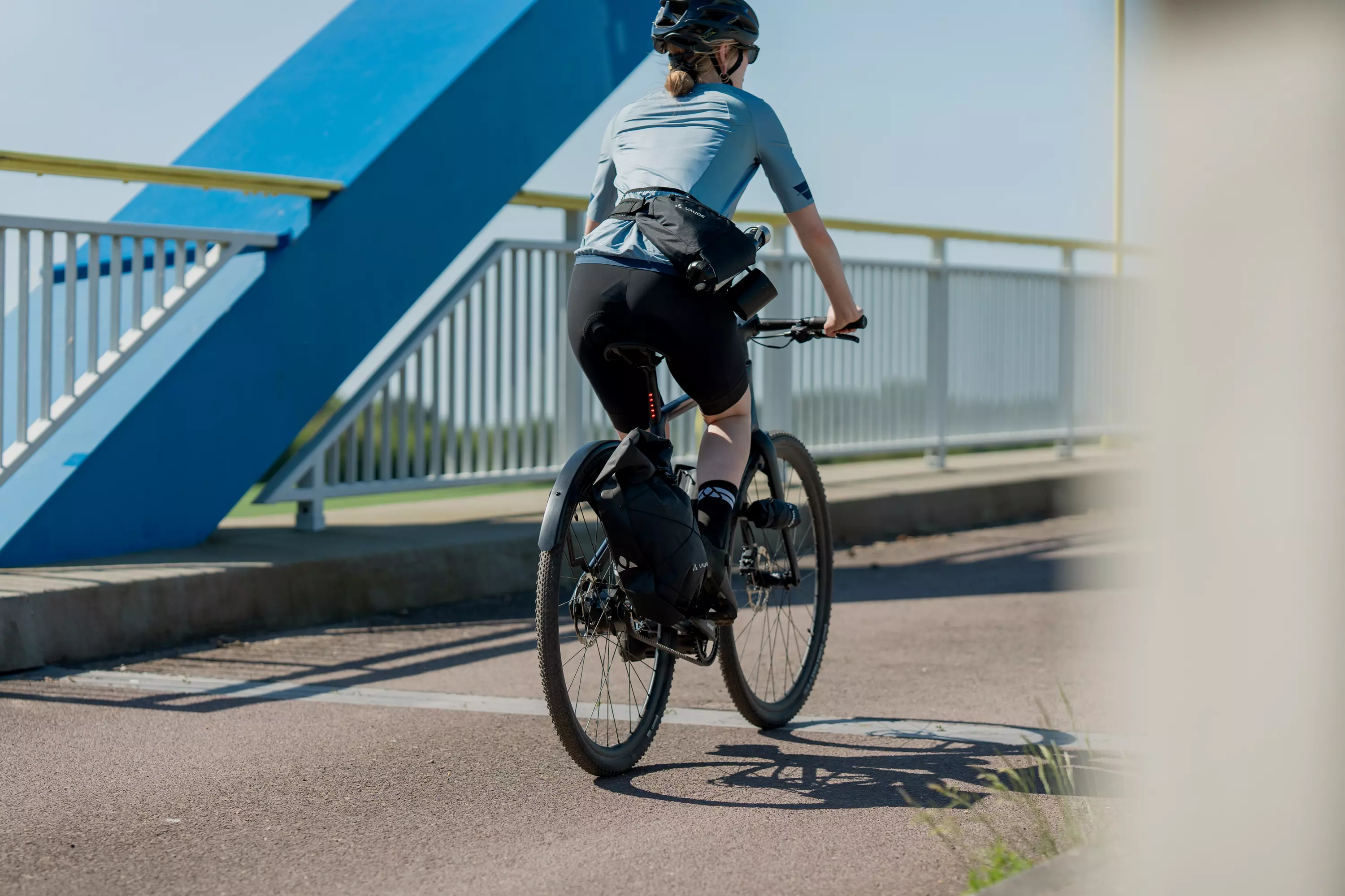 Junge Frau fährt mit ihrem Gravel E-Bike Waldwiesel in Rückansicht über eine Brücke